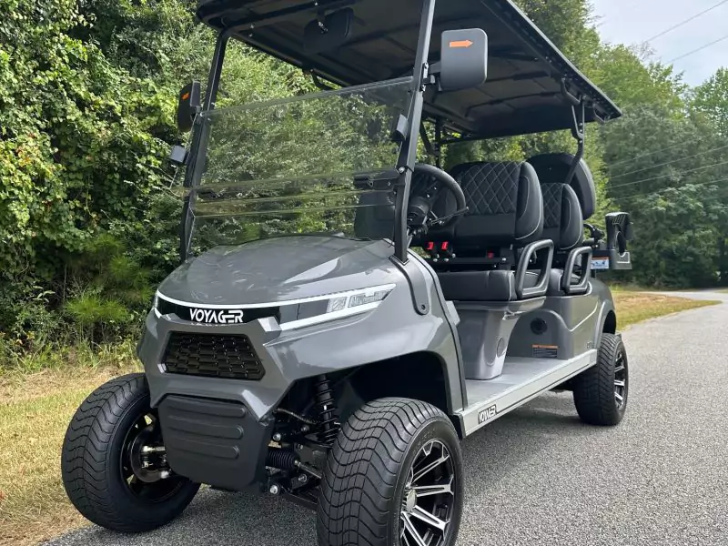 Gray Voyager golf cart with black diamond-stitched seats, folding windshield, and black alloy wheels parked on a road beside wooded greenery.