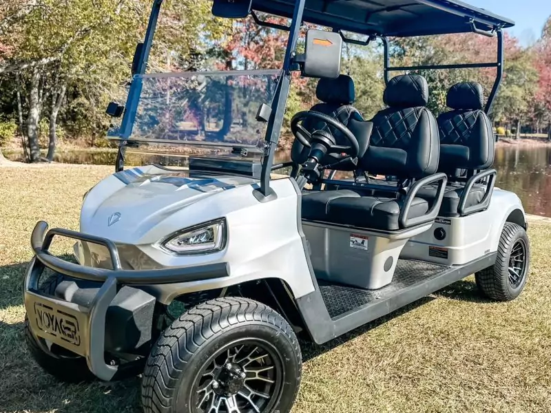 Silver Voyager golf cart with black diamond-stitched seats and alloy wheels parked near a pond with fall trees in the background.”
