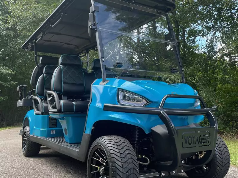 Blue 6-passenger Voyager golf cart featuring black diamond-stitched seats with blue piping, front brush guard, alloy wheels, and a clear folding windshield parked beside trees.