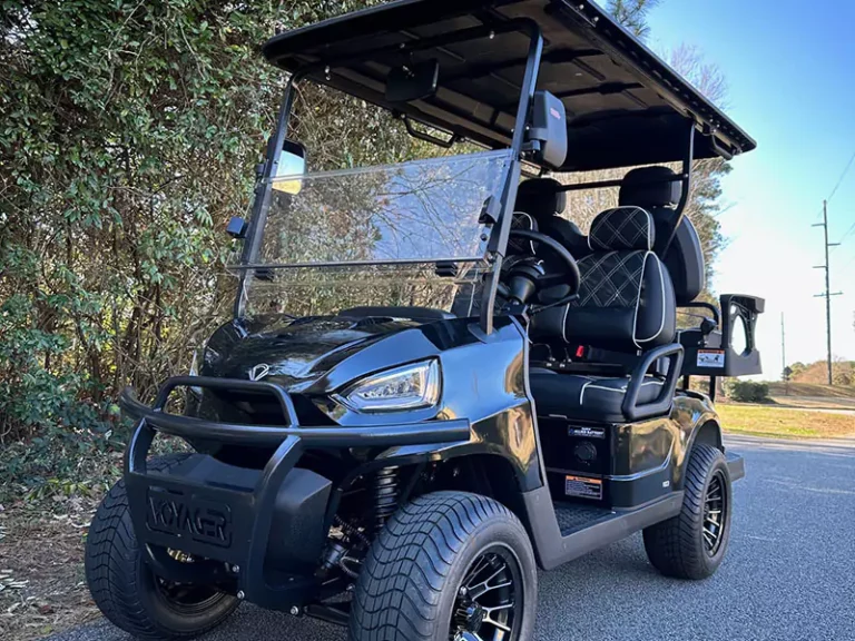 Black Voyager golf cart with diamond-stitched seats, lifted suspension, and chrome wheels parked on a roadside near wooded trees.