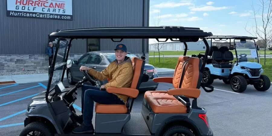 Man sitting in a gray golf cart with tan seats outside the Hurricane Golf Carts dealership. A second cart is parked nearby, showcasing available models at HurricaneCartSales.com.
