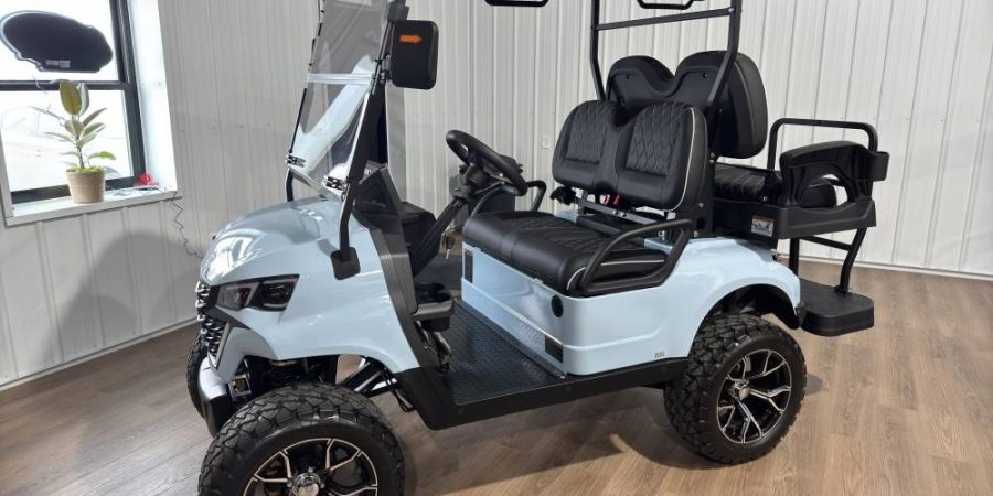 Light blue 4-passenger Breezy electric golf cart with black premium seats, rear flip deck, rugged off-road tires, and overhead canopy in a showroom setting.