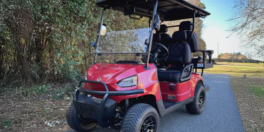 Red Voyager lifted golf cart with black seats and off-road wheels parked on a paved path near trees.